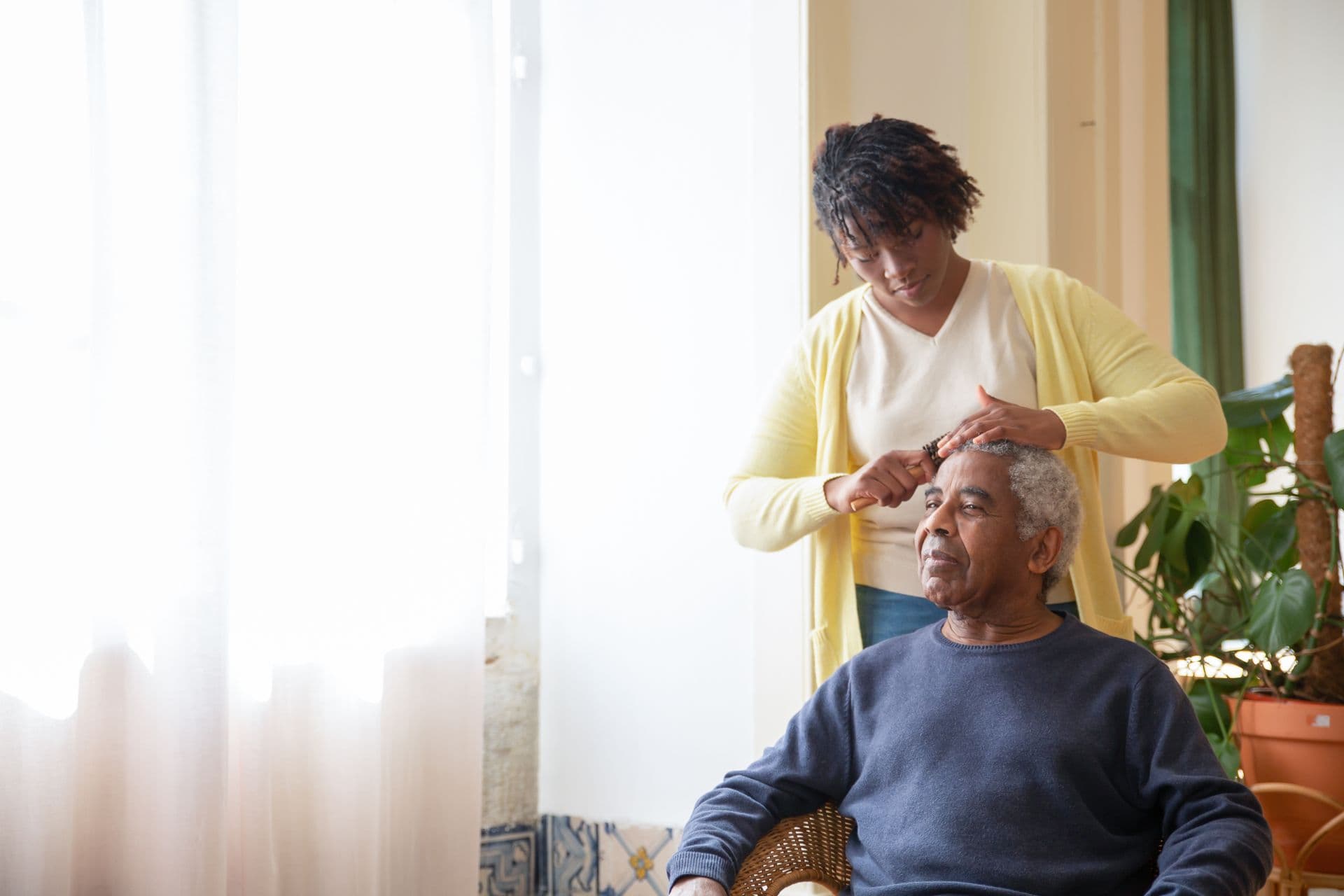 A carer gently brushing an older man’s hair by a sunlit window